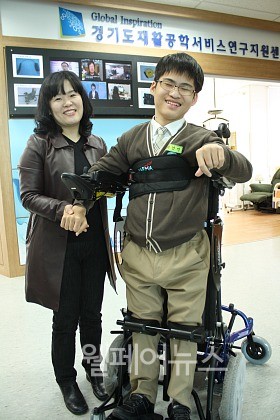 ▲ 학습용 맞춤 보조기구를 지원받은 박건민(오른쪽)군이 기립전동휠체어로 스스로 일어서는 모습에 어머니가 함께 웃고 있다.